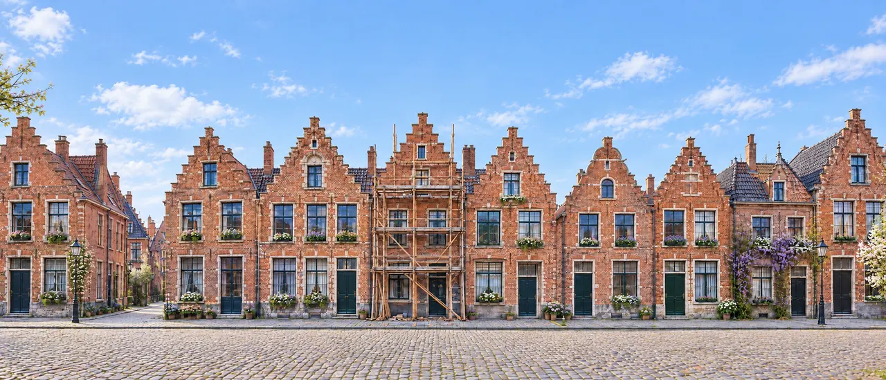Een Vlaamse straat met trapgevels en bloembakken onder een blauwe lucht. Eén huis staat in de stelling.